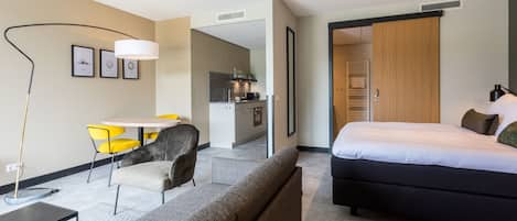 A modern hotel room with a large bed, a sofa, a dining area with yellow chairs, and a kitchenette.