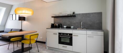 A modern kitchen with a dining area, featuring a round table, yellow chairs, a built-in oven, and a shelf with glasses.