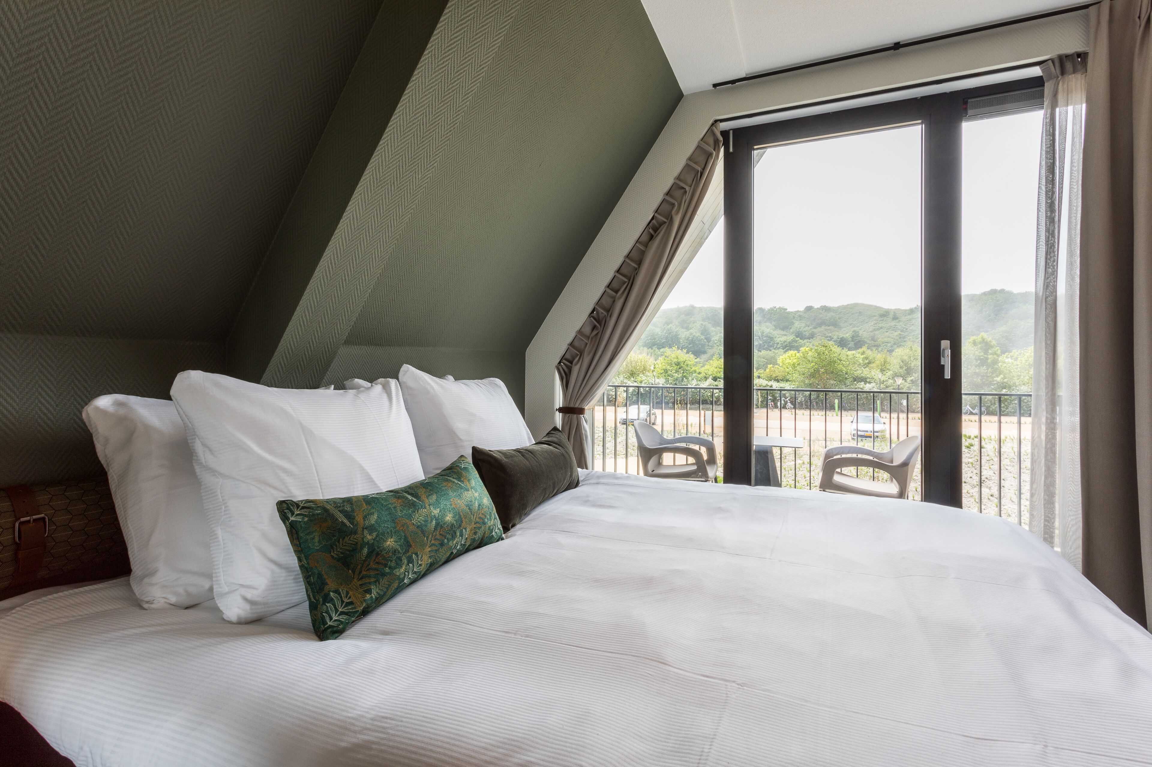 A hotel room with a large bed, a view of the outdoors through a sliding glass door, and a balcony with chairs.