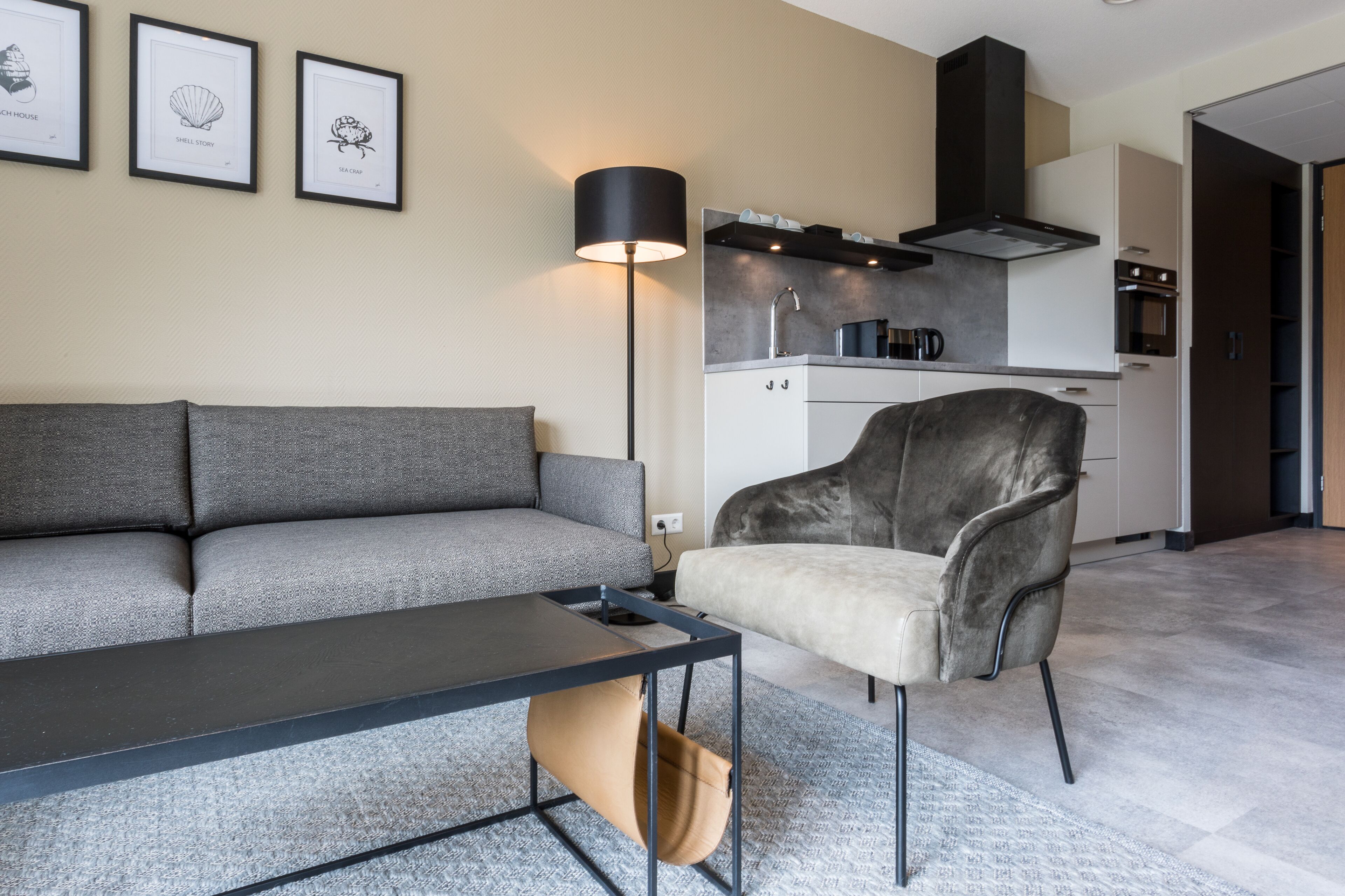 Una sala moderna con un sofá gris, una mesa de centro oscura y un sillón gris claro. Se observa un área de cocina con armarios blancos y una campana extractora negra. Tres cuadros enmarcados decoran la pared.