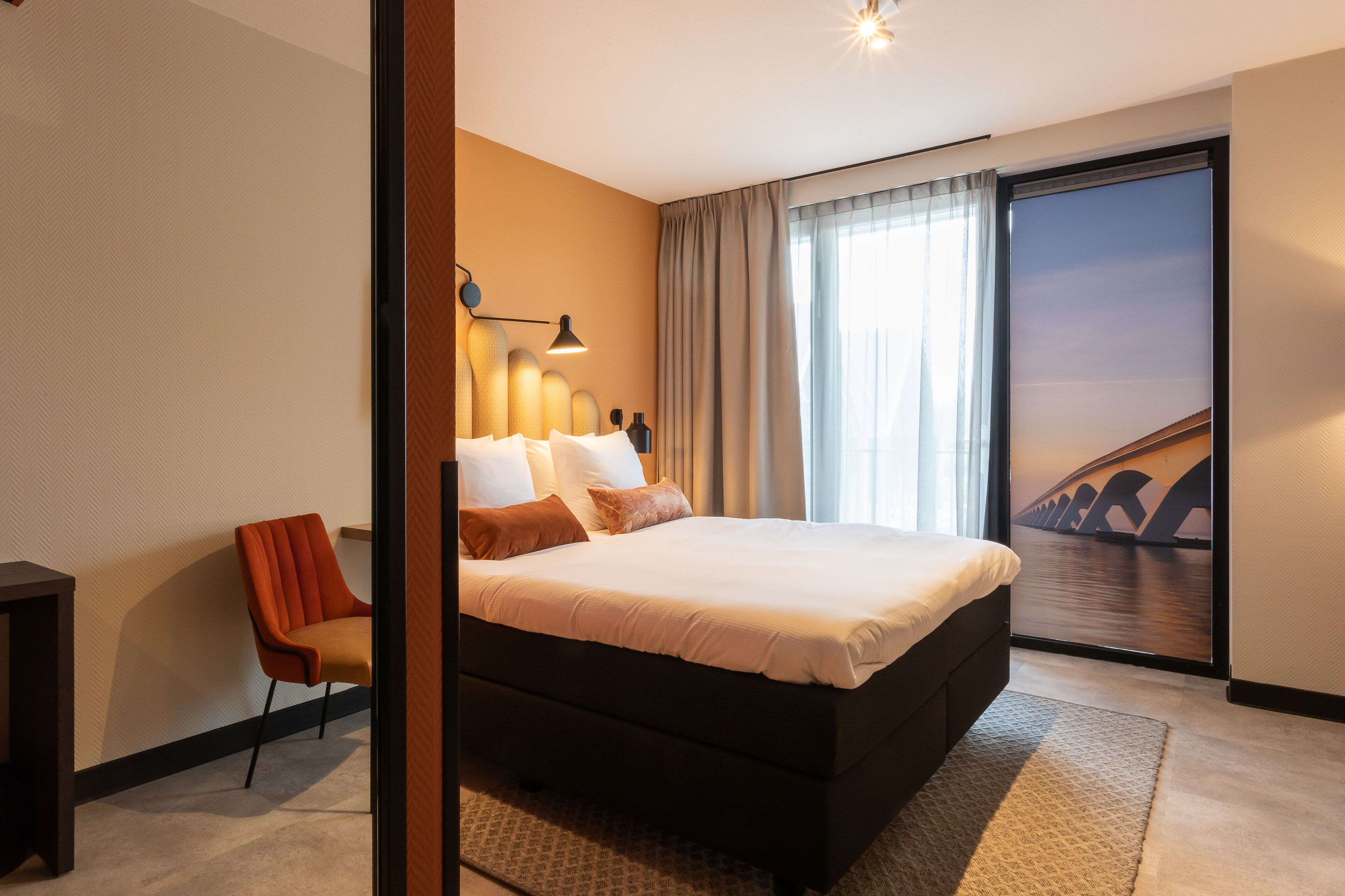 A modern hotel room with a large bed, a chair, a desk, and a large window with a view of a bridge.