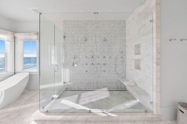 A modern bathroom with a glass-enclosed shower, a large bathtub, and a view of the ocean.