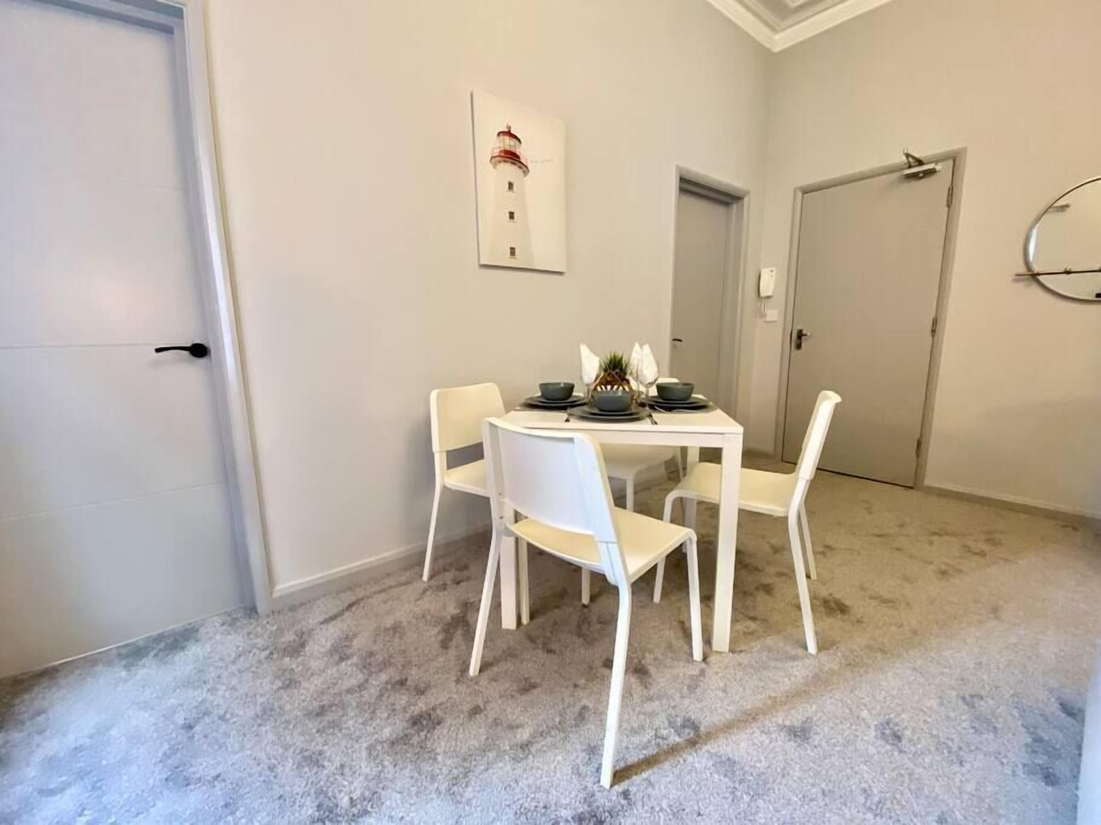 Un comedor con una mesa blanca y cuatro sillas, un cuadro en la pared, y piso alfombrado.