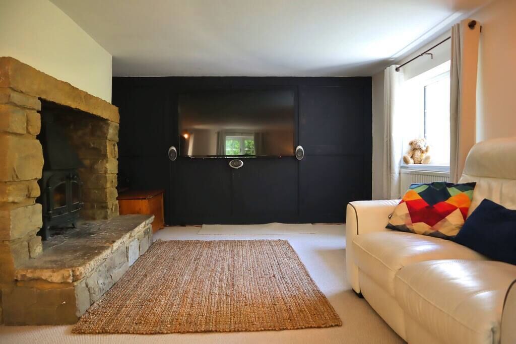 Uma sala de estar com lareira de pedra, parede escura, sofá branco com almofadas coloridas e janela com um urso de pelúcia no parapeito.