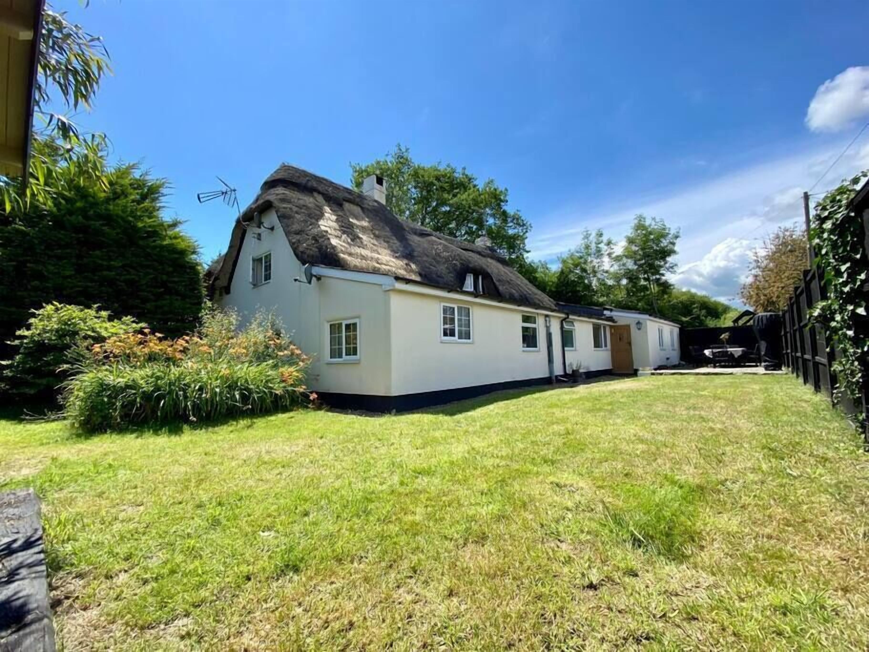 A white house with a thatched roof, surrounded by a well-maintained lawn and dense foliage.
