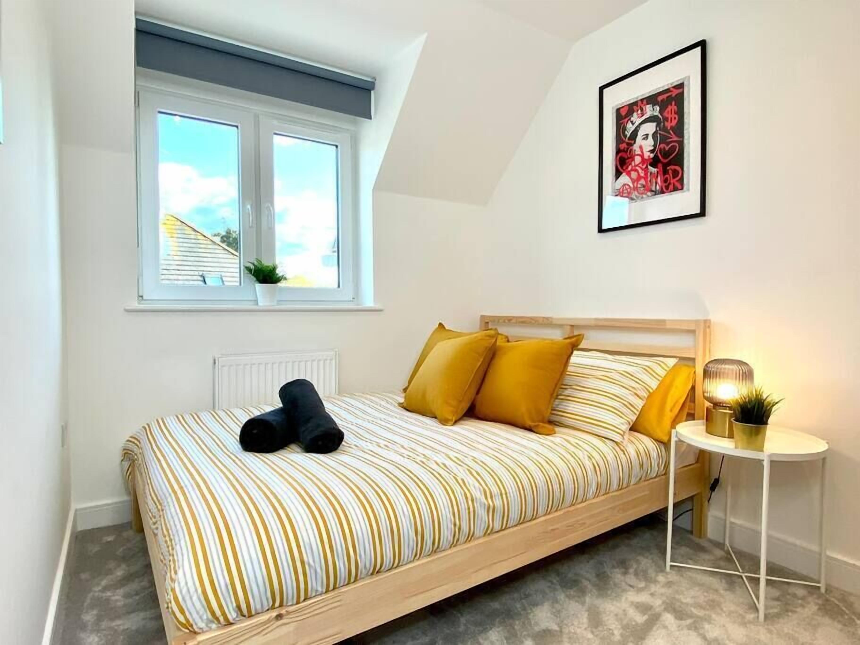 Un dormitorio con cama, ventana, radiador, una mesita con lámpara y un cuadro en la pared.