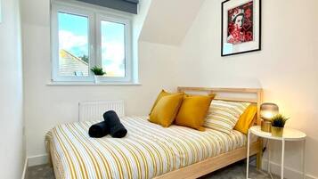 Un dormitorio con cama, ventana, radiador, una mesita con lámpara y un cuadro en la pared.