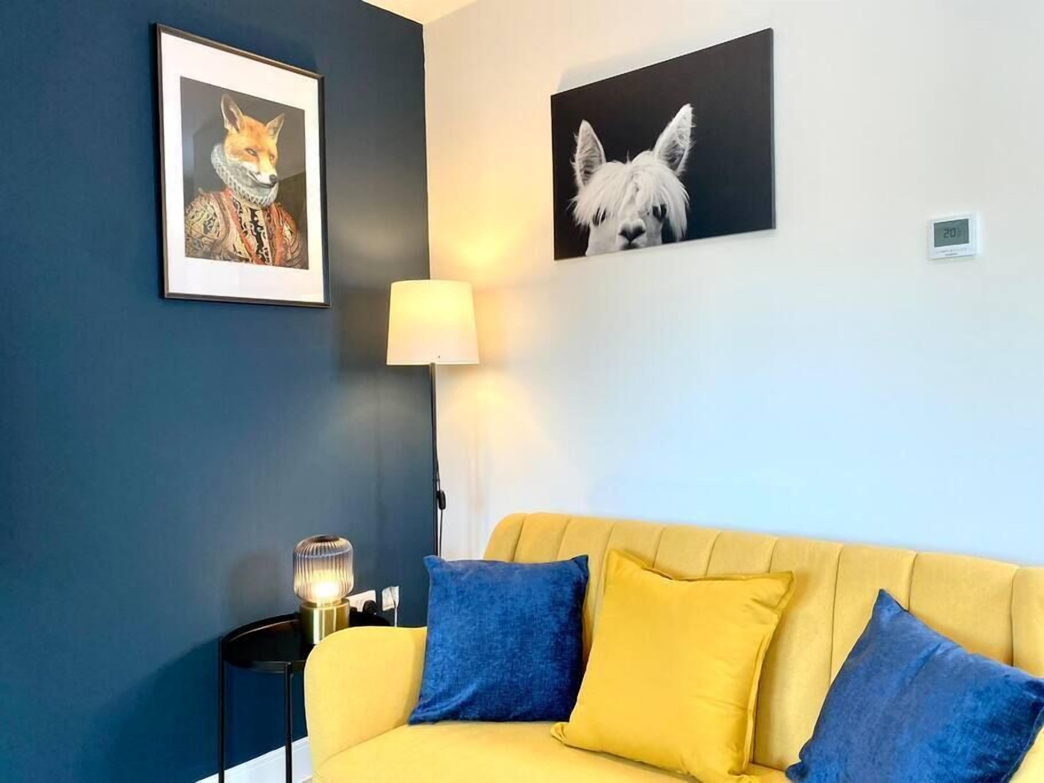 A room with a yellow sofa, two blue pillows, a framed fox painting, a framed dog painting, a lamp, and a thermostat on the wall.