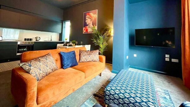 Ein modernes Wohnzimmer mit einer Couch, einem gemusterten Hocker, einem Fernseher und einer Küche im Hintergrund.