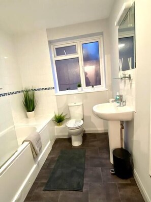 Une salle de bain avec des toilettes, un lavabo et une baignoire. Il y a une fenêtre au-dessus des toilettes et une petite plante sur le sol.