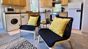 A compact kitchen with wooden cabinets, a microwave, a washing machine, and a refrigerator. There are two chairs with cushions and a small table with a kettle and a cup. - Trendy Apartment - 5 minute walk to the beach! (Bournemouth)