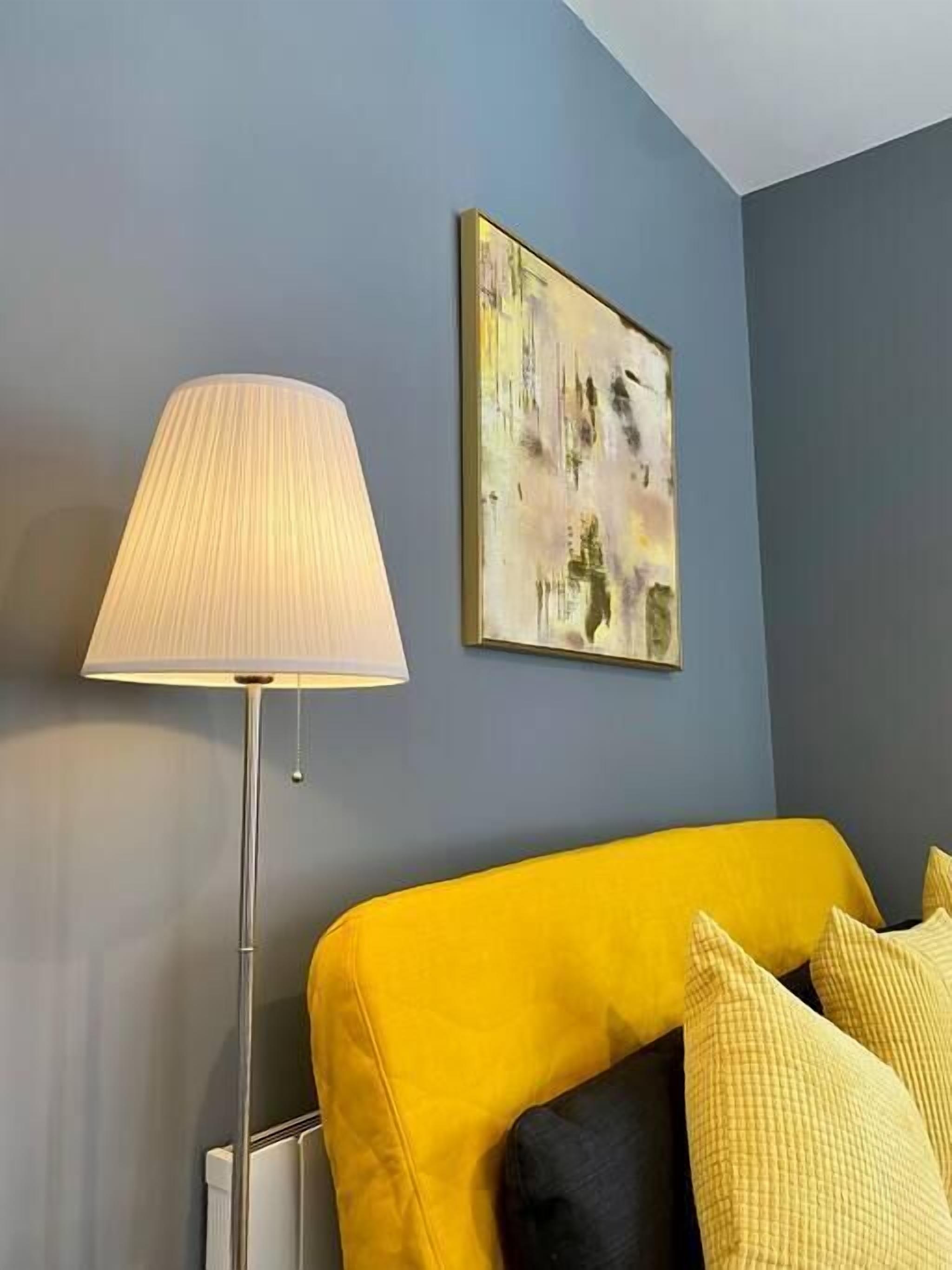 Une pièce avec un canapé jaune, un lampadaire et un tableau accroché au mur.