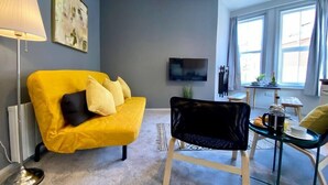 A living room with a yellow sofa, a black chair, a TV, and a dining area with a table and chairs. - Trendy Apartment - 5 minute walk to the beach! (Bournemouth)
