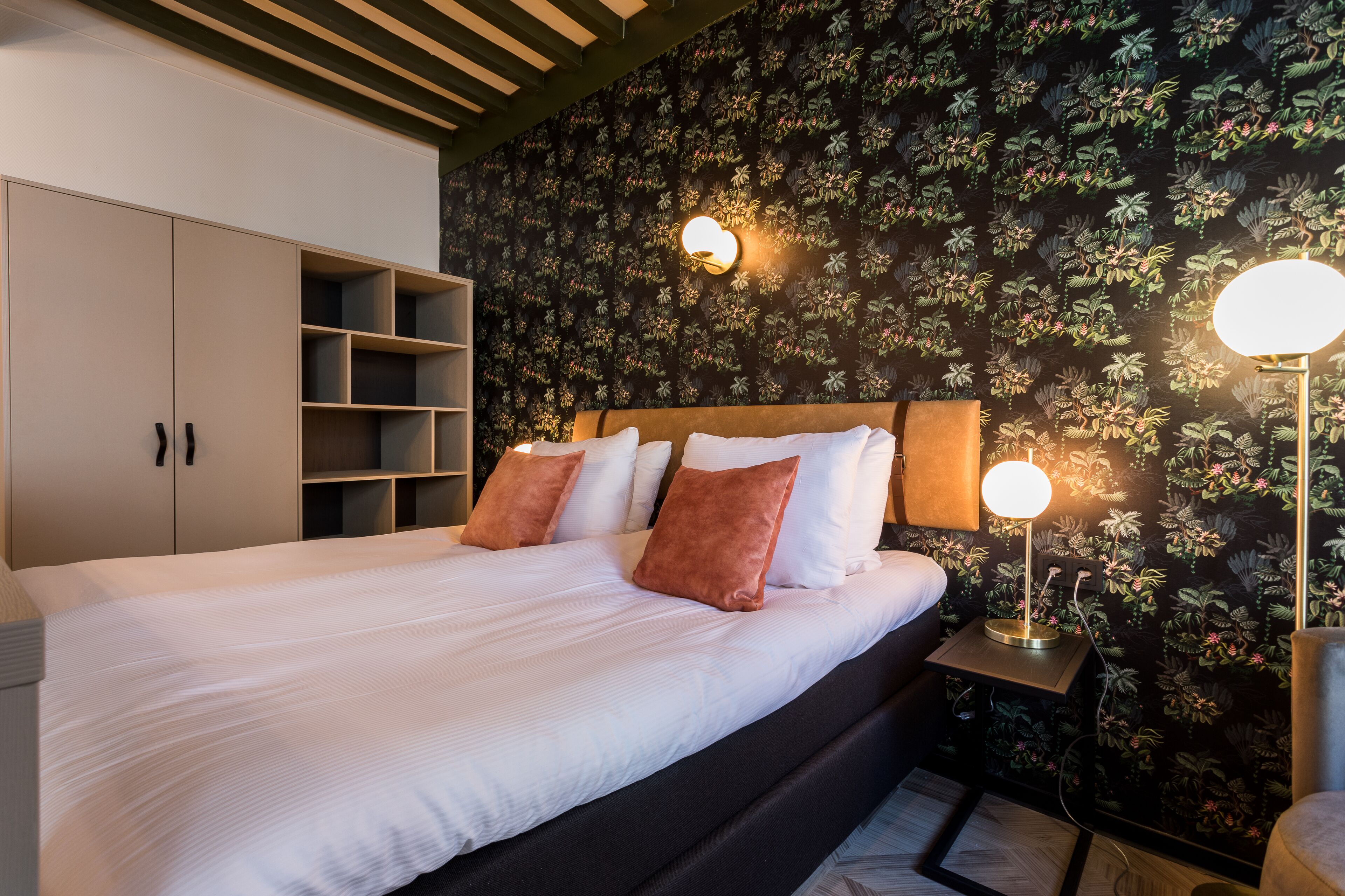 Un lit bien fait, avec des draps blancs et deux oreillers marron, une tête de lit en bois et un mur recouvert d’un papier peint à motifs floraux.