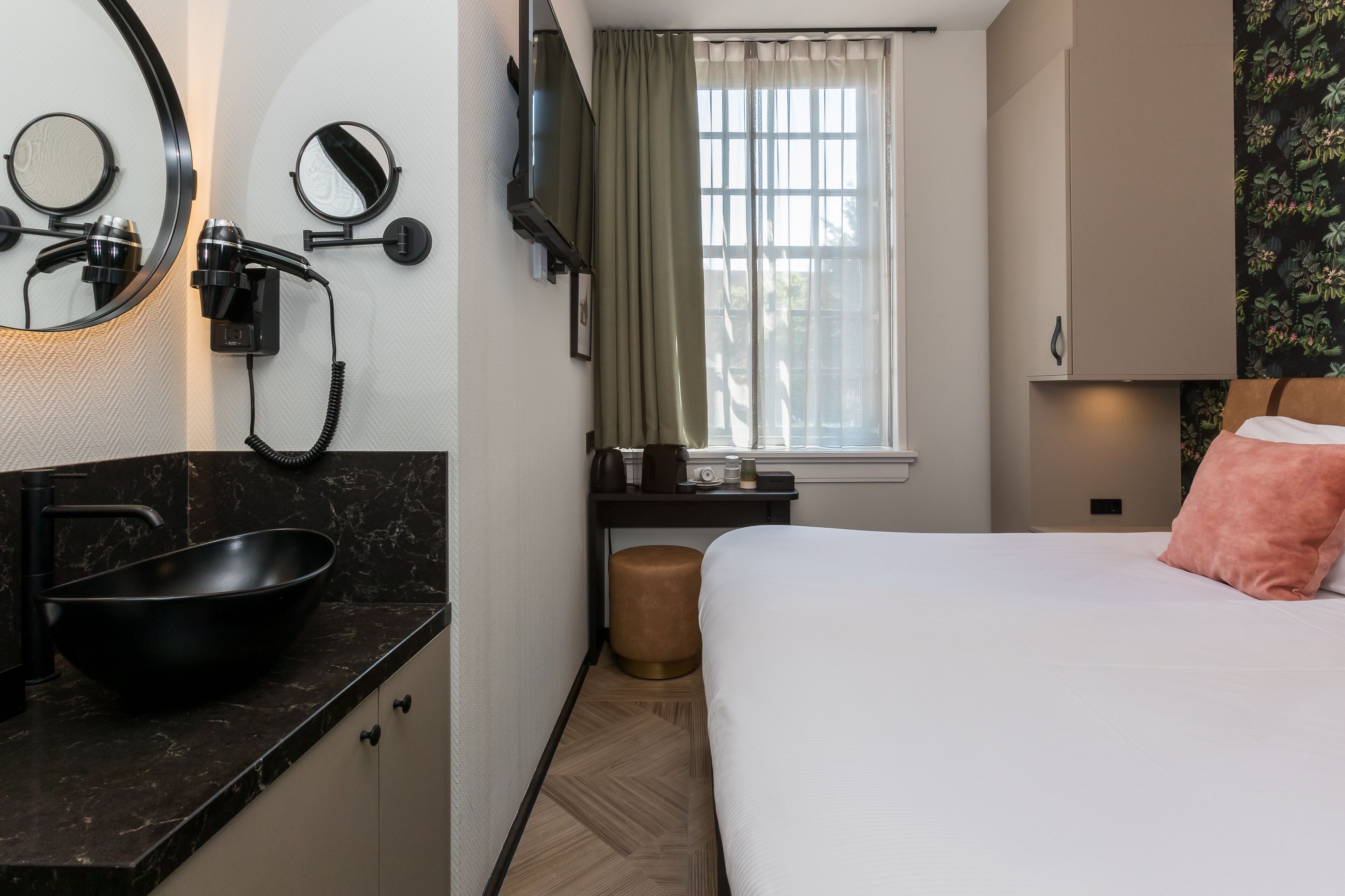Camera d'albergo con lavandino in marmo nero, specchio rotondo, asciugacapelli, televisione, finestra con tende e un letto con cuscino rosa, più uno sgabello.