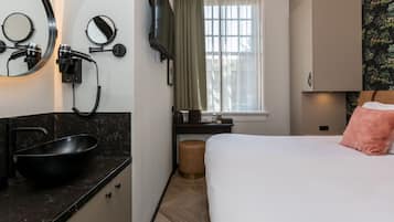Camera d'albergo con lavandino in marmo nero, specchio rotondo, asciugacapelli, televisione, finestra con tende e un letto con cuscino rosa, più uno sgabello.