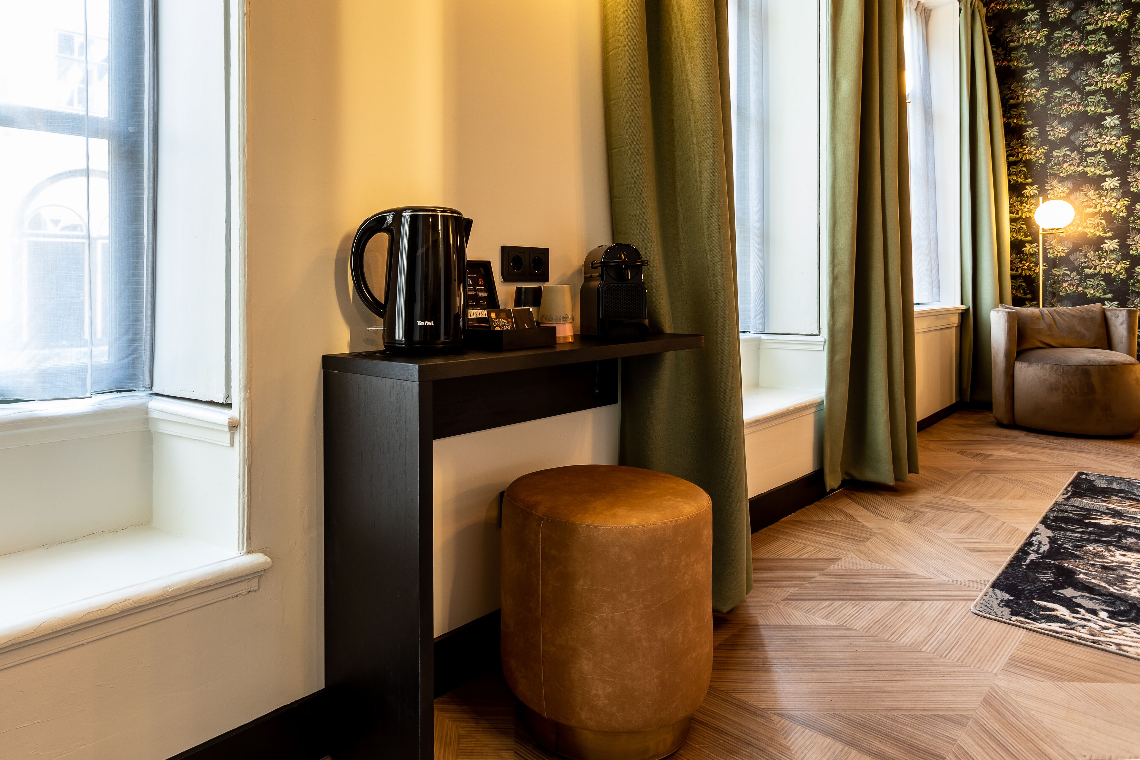 A room with a black console table, a brown ottoman, a black coffee maker, and a lamp.