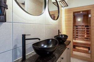 Un baño moderno con dos lavamanos negros, un grifo negro y una sauna de madera visible a través de una puerta de cristal.