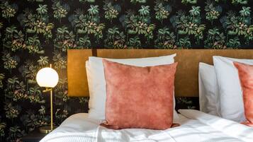A neatly made bed with white and pink pillows, a round table lamp, and a wall with a leafy pattern.