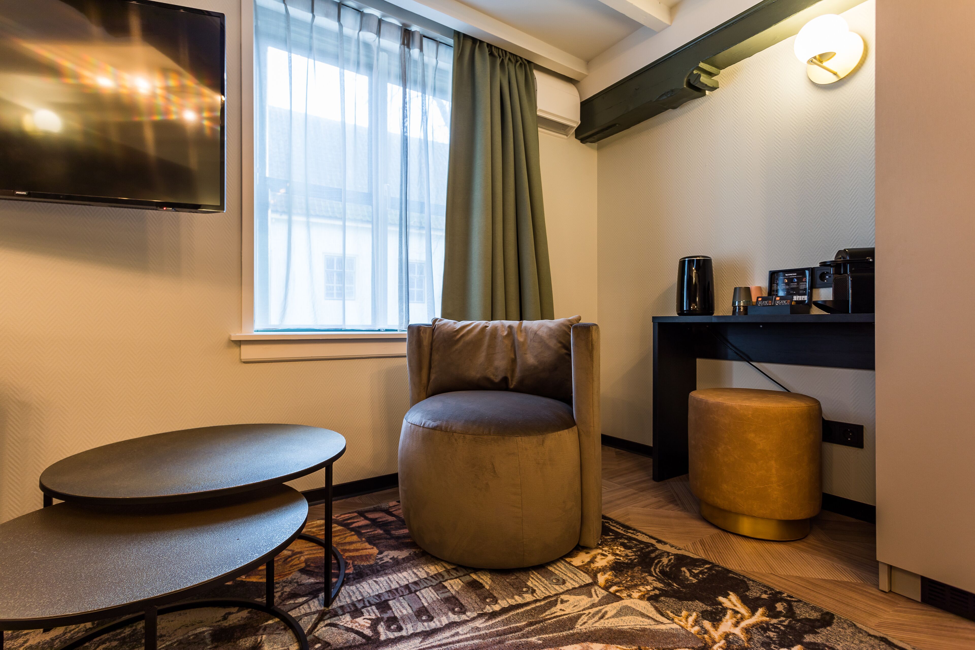 A modern hotel room with a flat-screen TV, a round ottoman, a small table, a desk with a coffee machine, and a window with curtains.