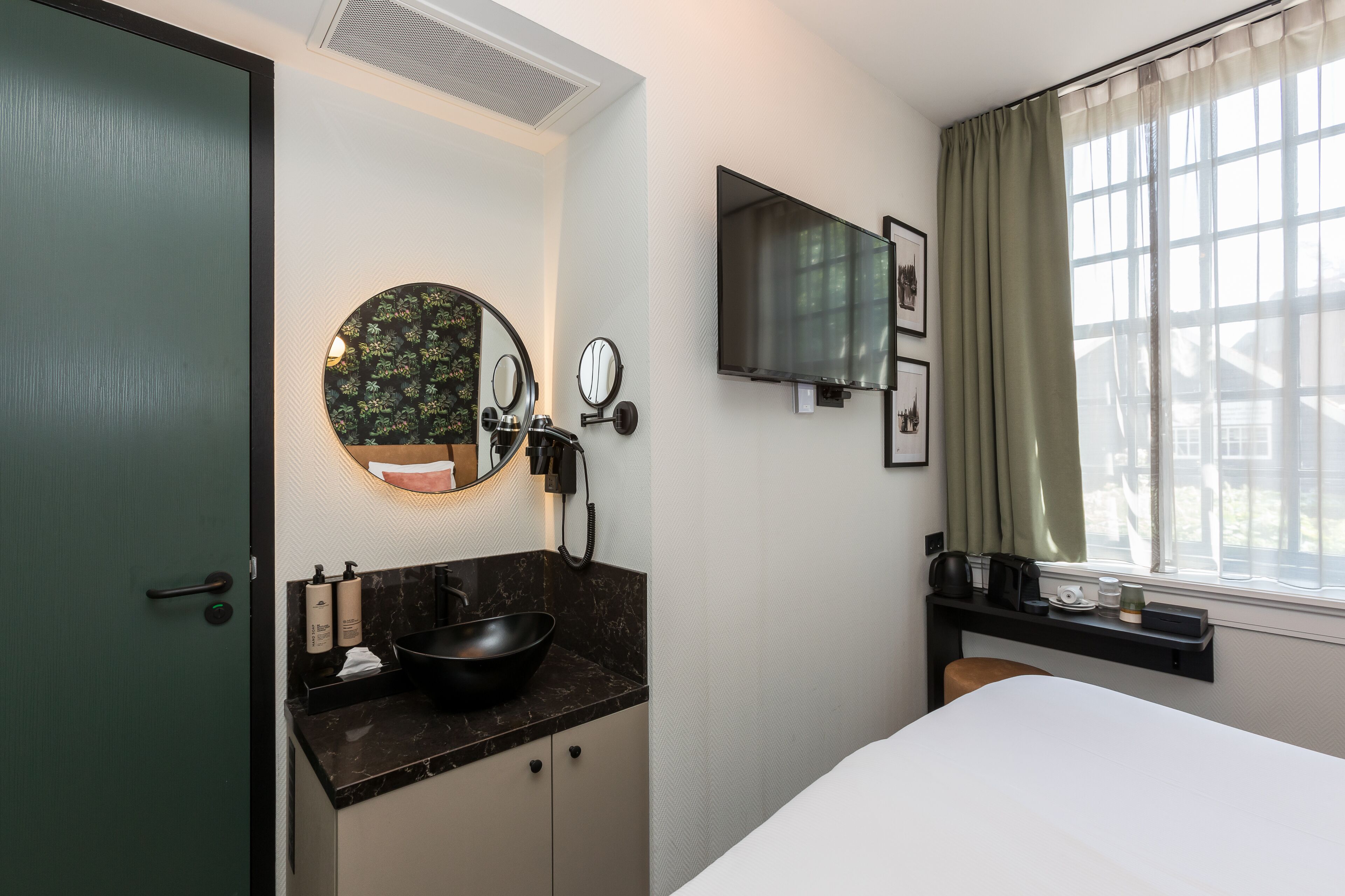 Habitación de hotel con cama, lavamanos de color negro, espejo redondo, televisor de pantalla plana montado en la pared y ventana con cortinas.