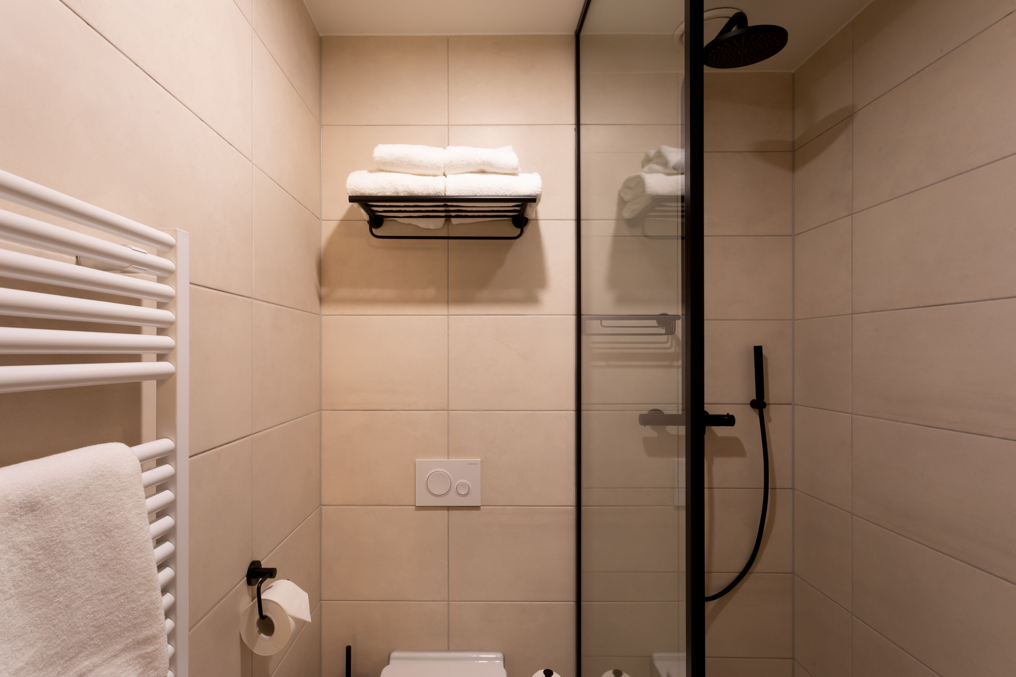 A modern bathroom with a glass shower enclosure, a towel rack with folded towels, a toilet paper holder, and a wall-mounted light switch.