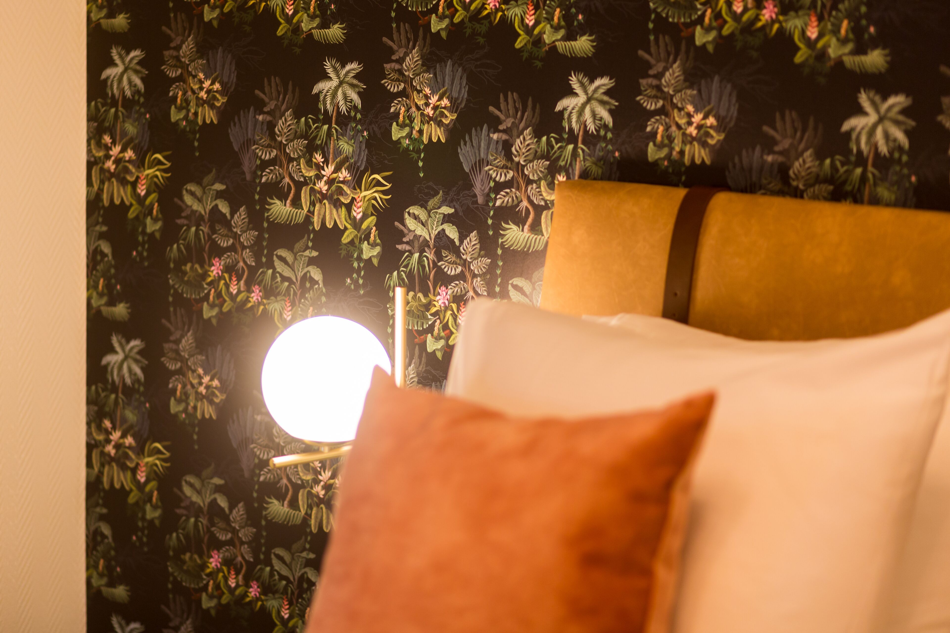 Une pièce avec un papier peint à motifs floraux, un lit avec des coussins blancs et bruns, et un luminaire rond fixé au mur.