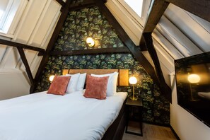A bedroom with a sloped ceiling, a bed with white bedding and two brown pillows, a nightstand with a lamp, and a wall with a floral wallpaper pattern.