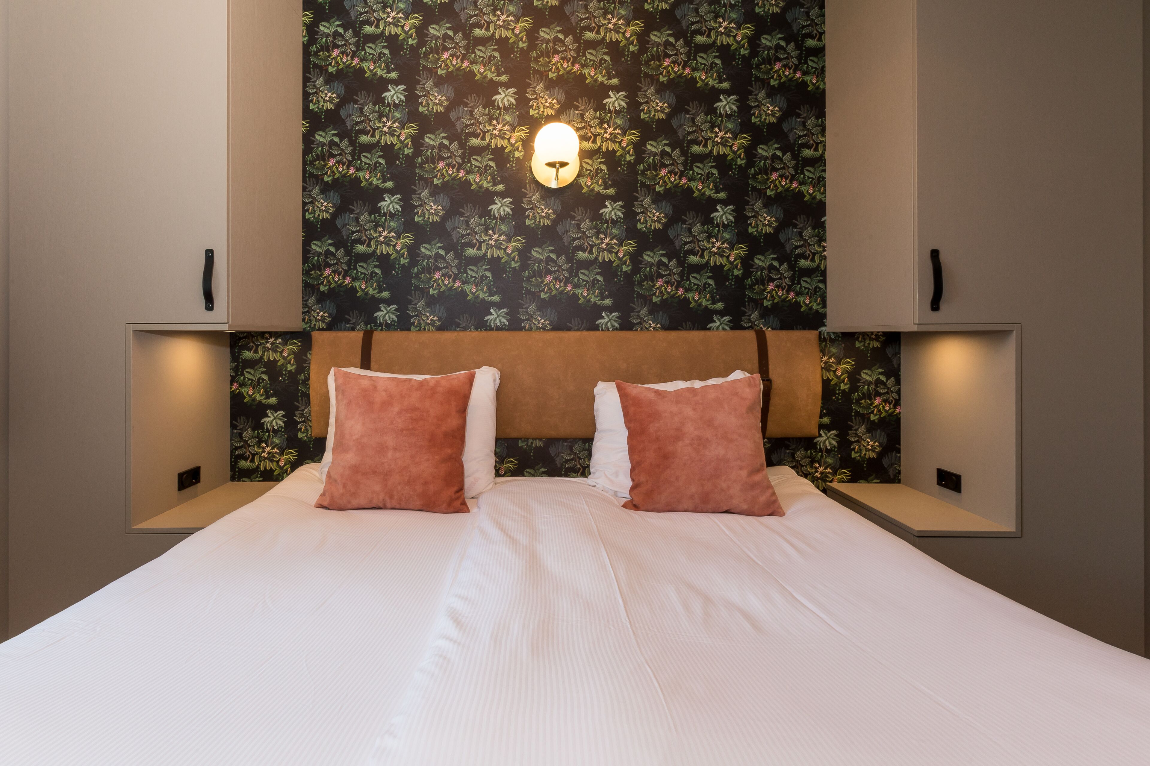 Quarto de hotel com uma cama grande, duas mesas de cabeceira, uma luminária fixada na parede e um papel de parede com padrão floral.