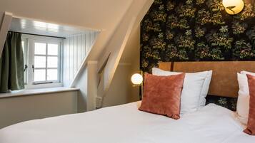 Un dormitorio con techo inclinado, una ventana con cortinas, una cama con ropa de cama blanca y dos almohadas, y una pared con estampado de flores.