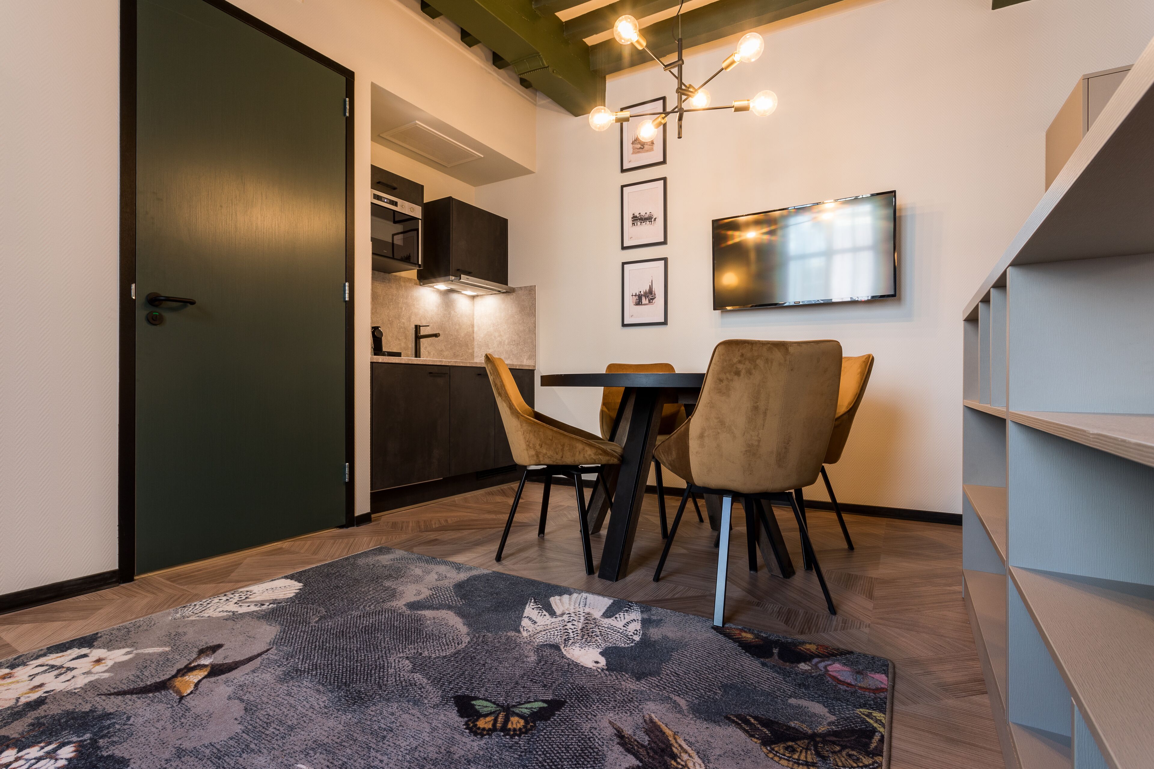 Un espace repas moderne avec une porte vert foncé, une cuisine en arrière-plan, une table à manger avec des chaises, un tapis à motifs et un téléviseur fixé au mur.