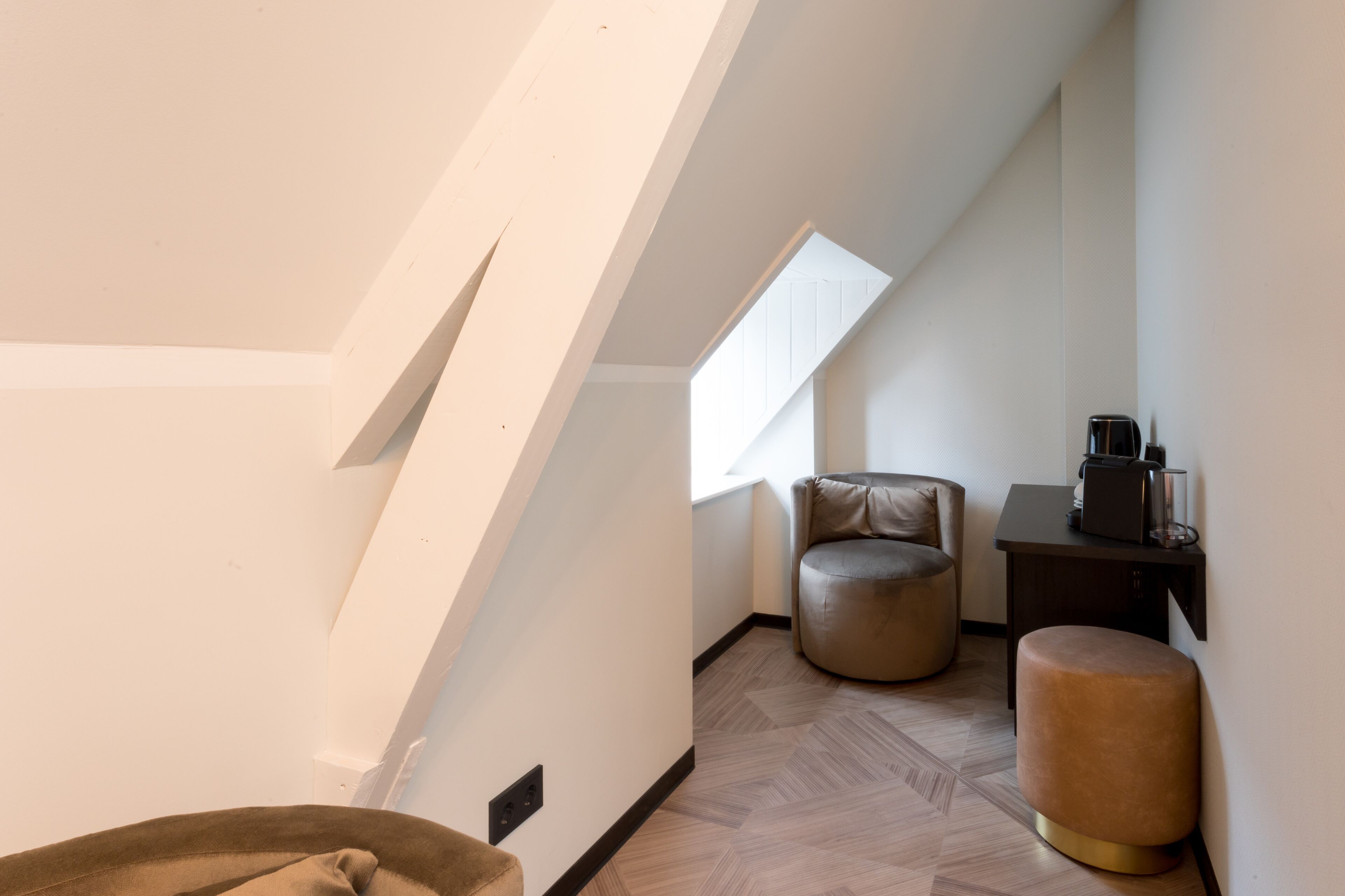 Una habitación estrecha de techos altos y con inclinación, una ventana, un sillón redondo de cuero, un pequeño reposapiés y una mesita de noche de madera oscura con una cafetera.