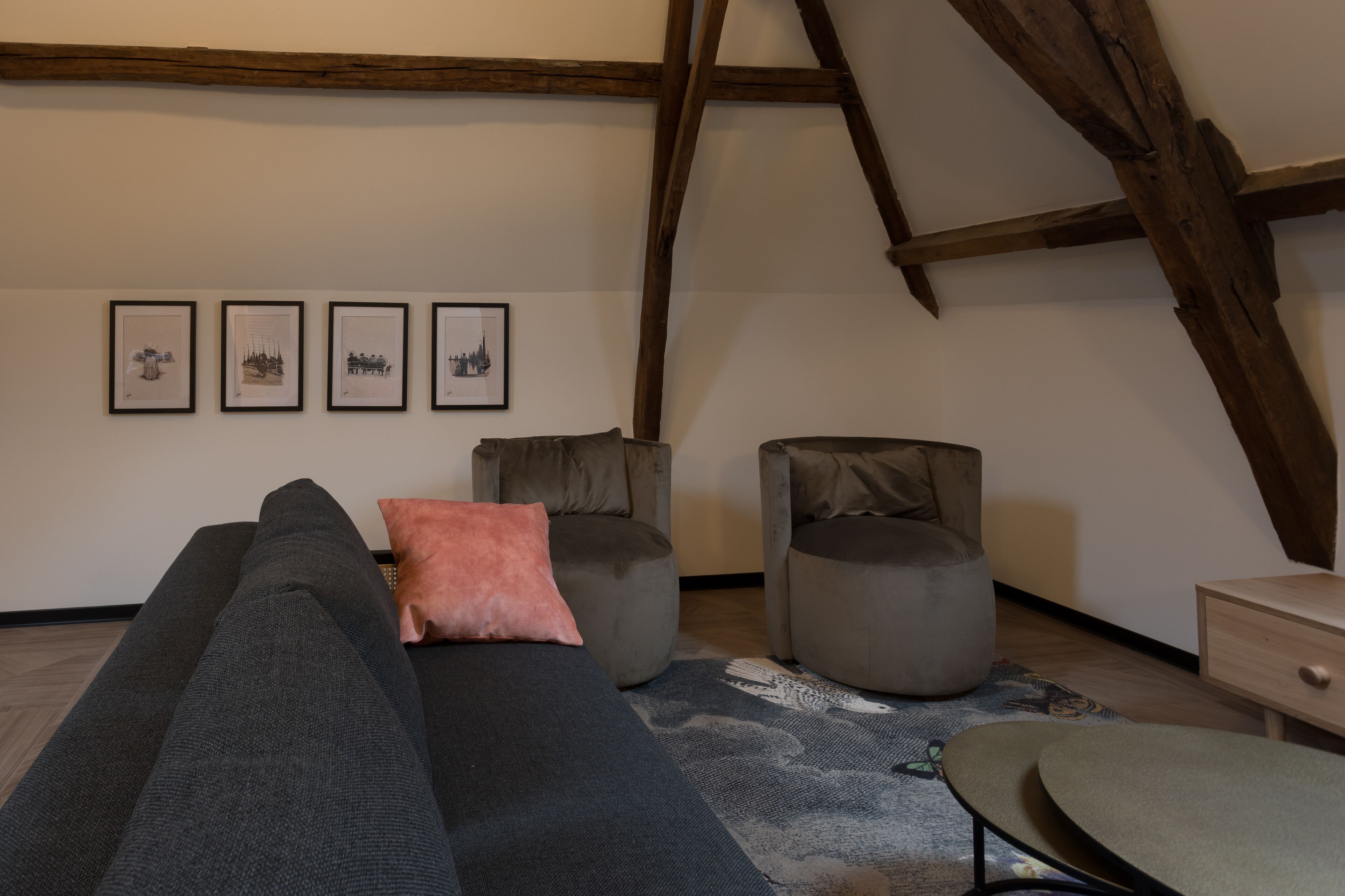 Un dormitorio estilo loft con un sofá gris, dos sillones grises, una mesita redonda y una escalera de madera. Hay cuatro cuadros enmarcados en la pared.