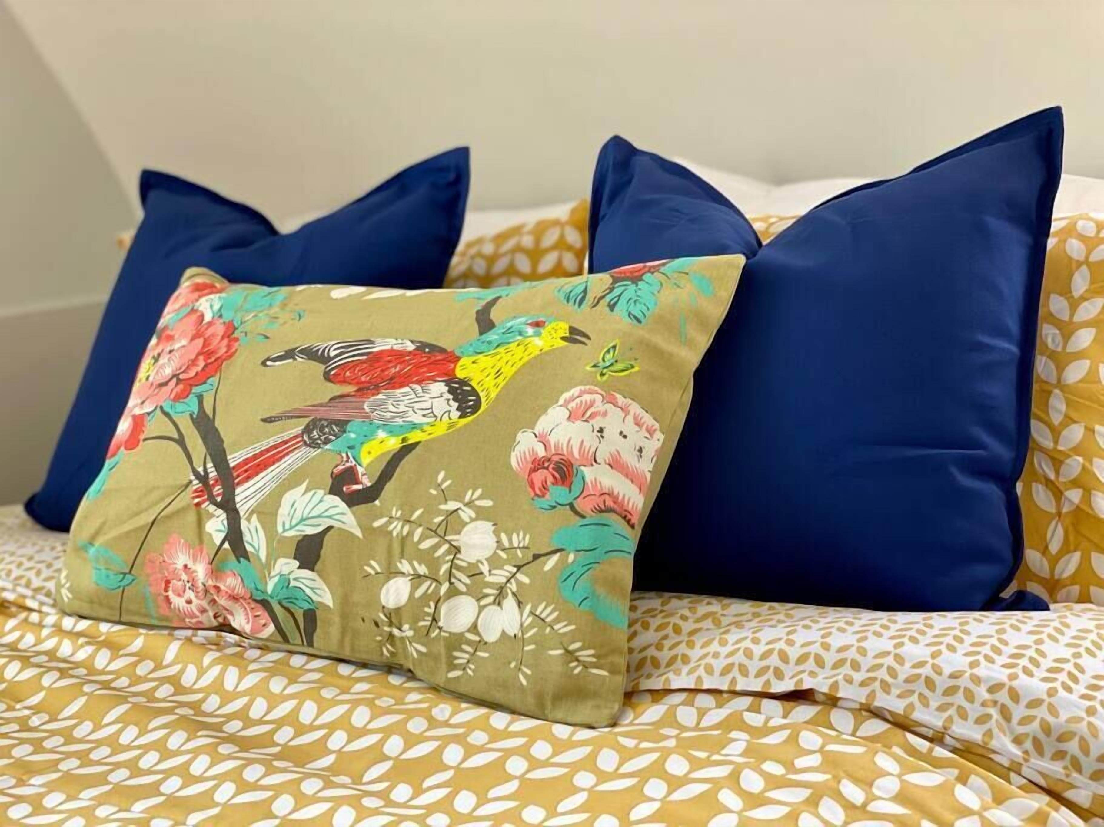A bed with a floral-patterned bedspread, a colorful pillow with a bird and flowers, and two solid blue pillows.