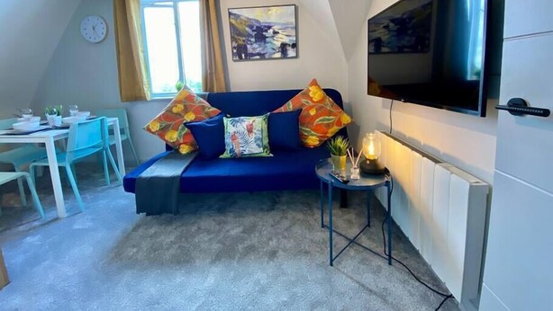 Un espacio habitable compacto con un sofá azul, una mesa de comedor con sillas, un televisor montado en la pared y una ventana con cortinas.