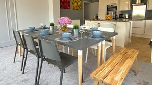 A modern dining area with a grey table set for six, surrounded by black chairs. A wooden bench is placed in front of the table. The room features a grey accent wall with fruit paintings, a white kitchen with white cabinets, and a hanging pendant light above the table. - Beautiful 4 Bedroom house -5 min walk to the beach (Bournemouth)