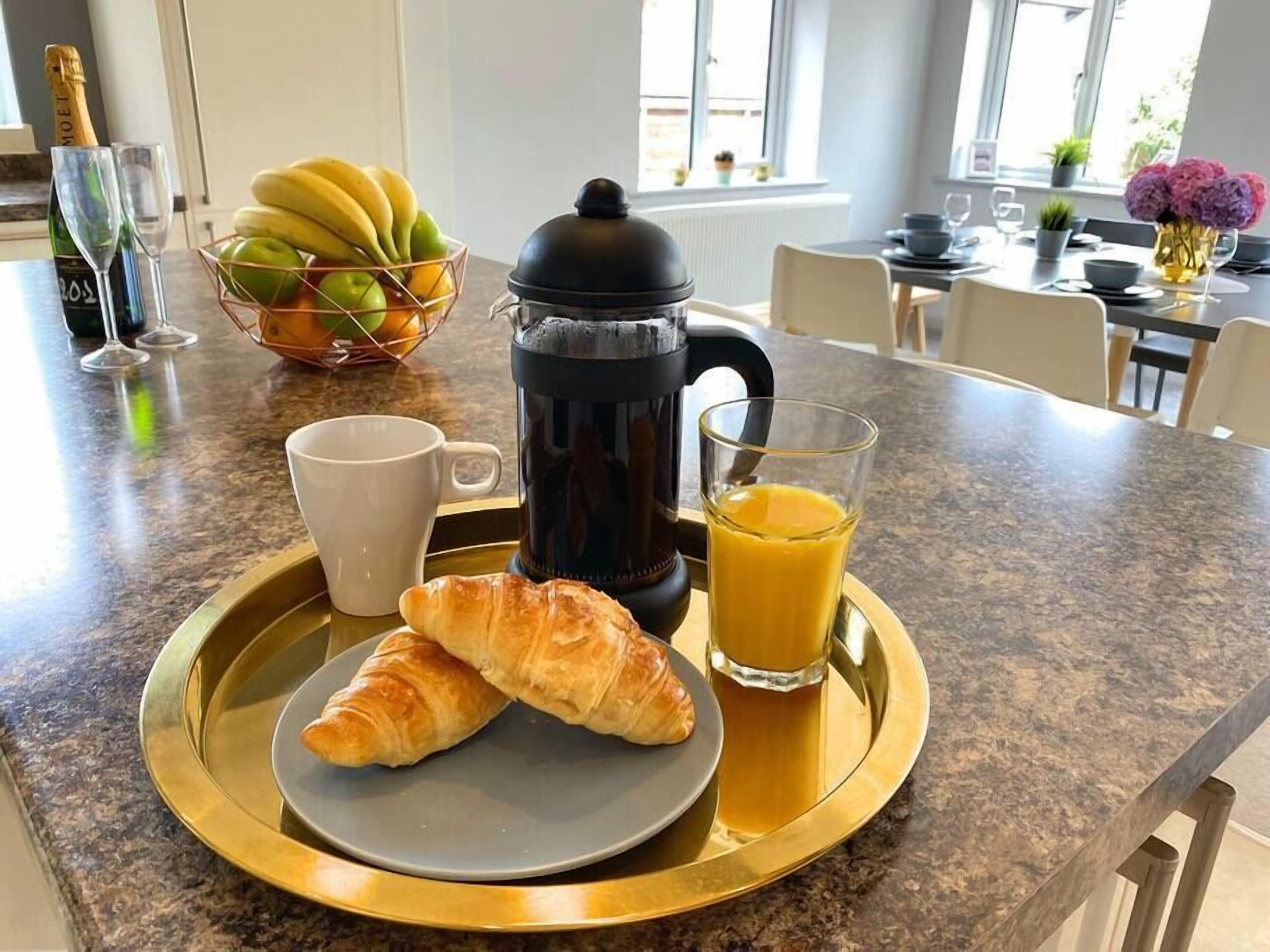 A breakfast tray with croissants, a coffee pot, a glass of orange juice, and a bowl of fruit on a kitchen counter.