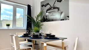 Un comedor con una mesa preparada para cuatro personas, sillas blancas, una planta en maceta y una ventana con vistas a casas.
