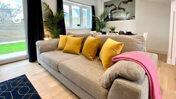 Una sala de estar moderna con un sofá gris, cojines amarillos, una manta rosa y un comedor al fondo.