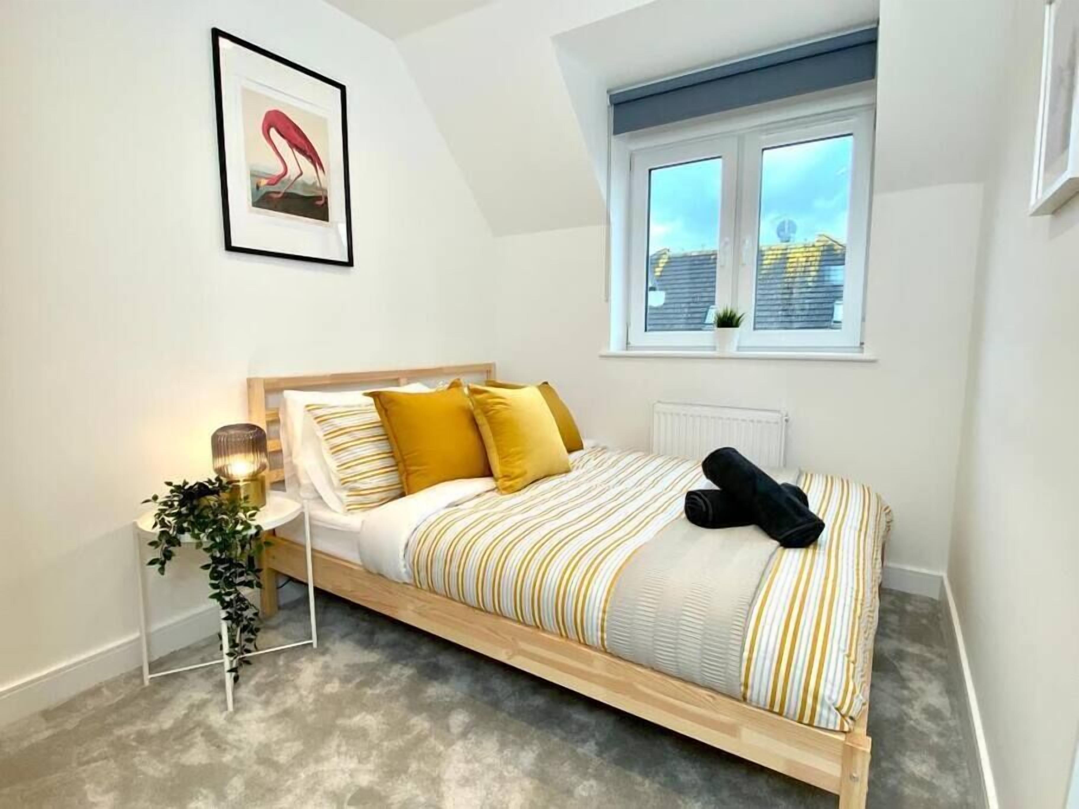 Un dormitorio con cama, mesita de noche, una planta, ventana con vistas y un cuadro en la pared.