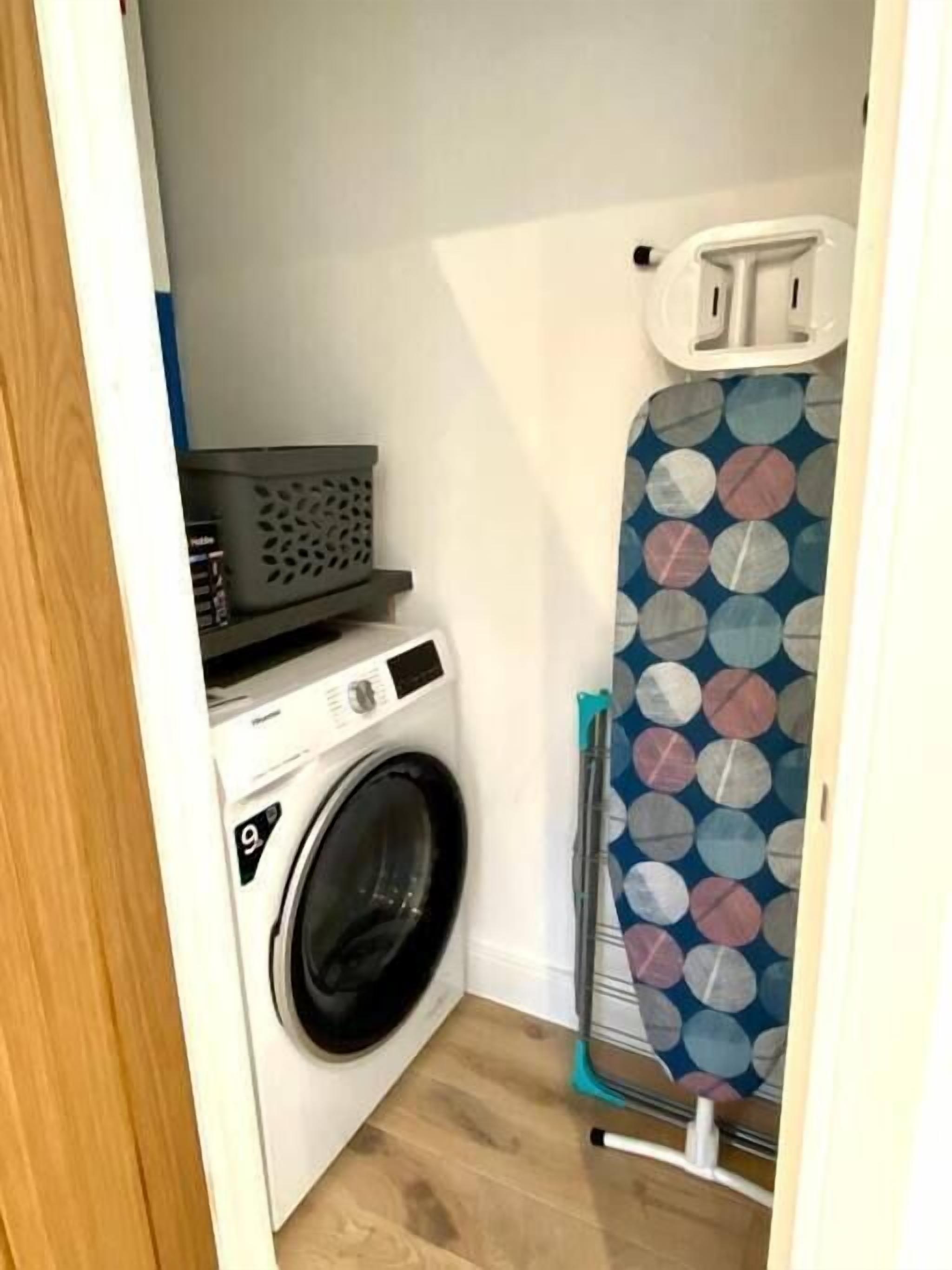 A compact laundry area with a front-loading washing machine, a drying rack, and a wall-mounted ironing board.