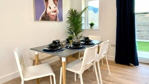 A dining area with a table set for four, white chairs, a potted plant, and a large painting of a cow on the wall.