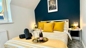 A bedroom with a bed, a bedside table, a nightstand, a framed picture on the wall, and a bottle of champagne on the bed.