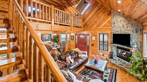 A log cabin interior with a stone fireplace, wooden staircase, and a cozy living area with a leather sofa and a television. - Super Views, Flat roads, Hot Tub (Gatlinburg)