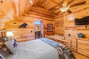 Un dormitorio acogedor tipo cabaña de madera con cama, escritorio, silla, ventilador de techo, televisor y ventana con vistas al exterior.