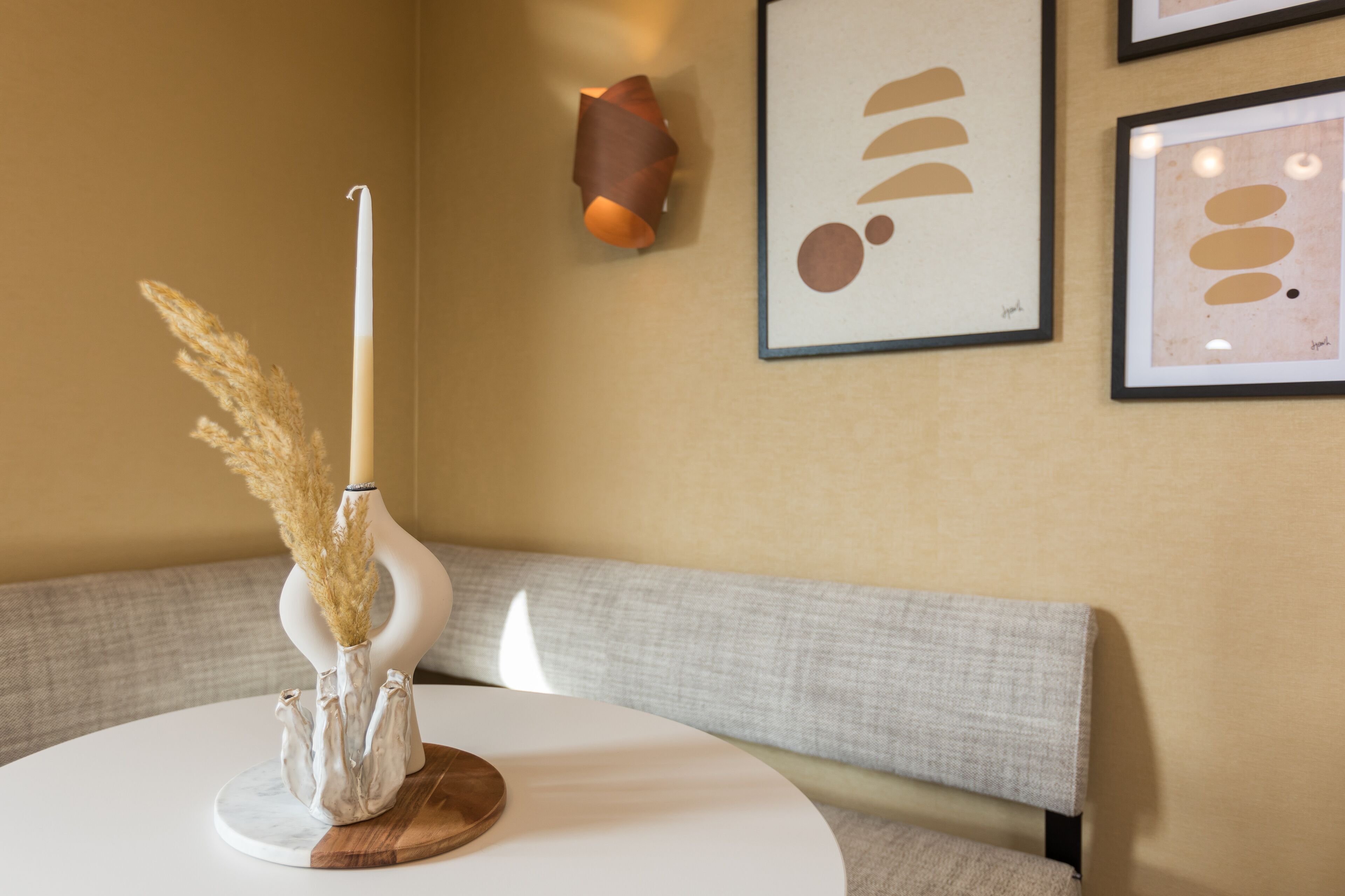 A round table with a white vase containing pampas grass and a candle, a gray sofa, and abstract art on the wall.