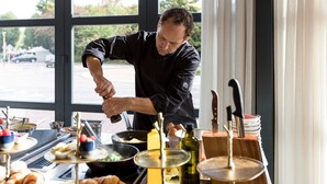 A chef preparing food in a kitchen with large windows, a view of the street outside, and various cooking utensils and ingredients on the counter.