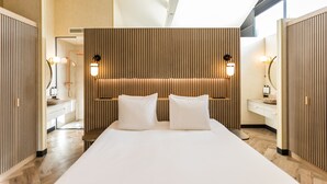 A modern hotel room with a large bed, wooden panel wall, bedside tables with lamps, and a bathroom visible through a glass door.