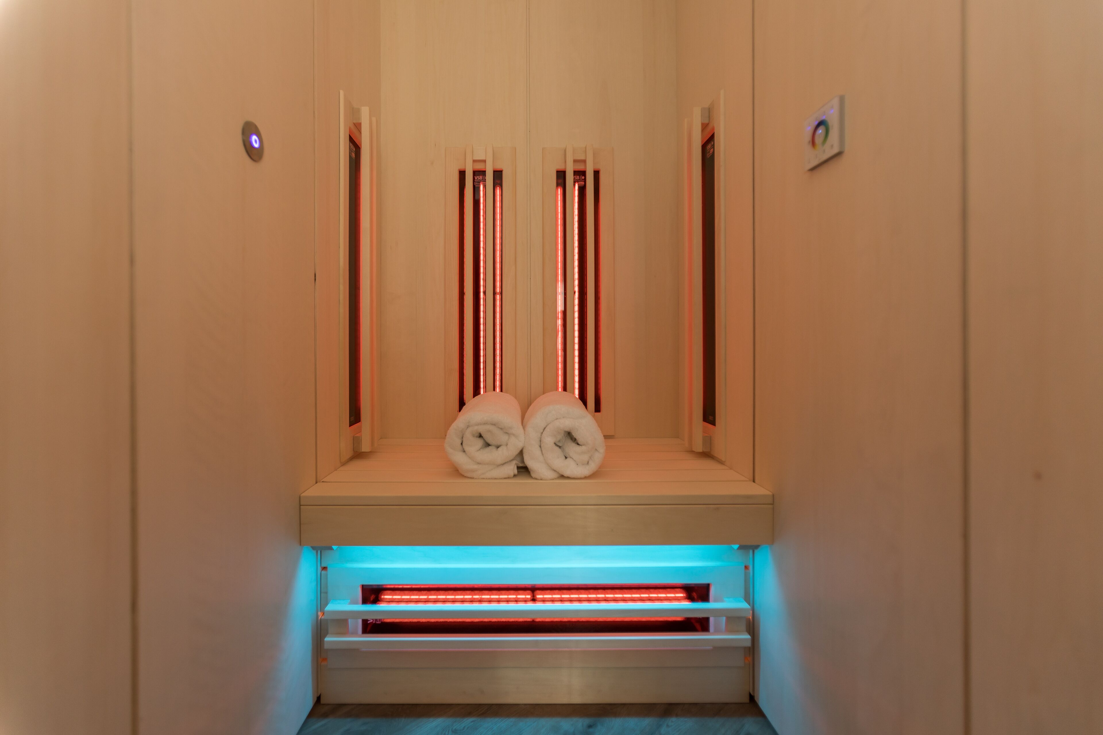 Una sauna con dos toallas sobre un banco, una luz azul en la parte inferior y un panel de control en la pared.