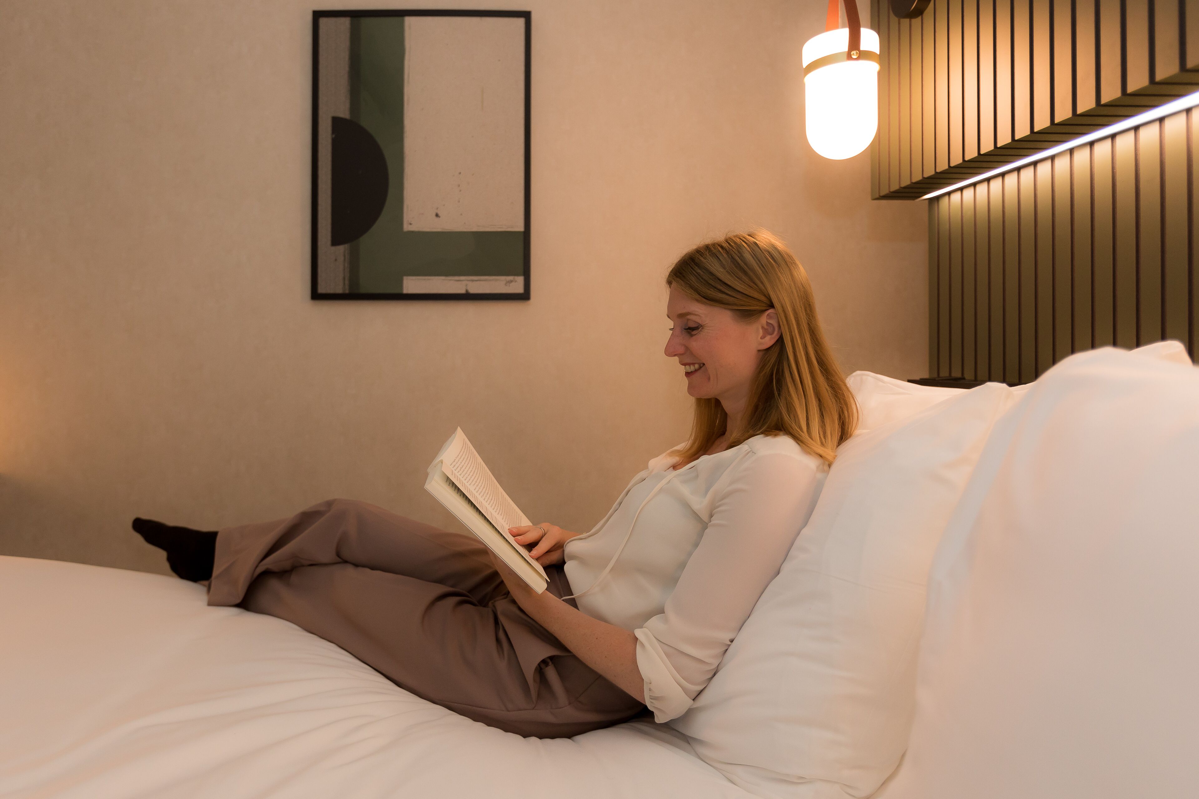 A person sitting on a bed in a room with a framed artwork on the wall and a wall-mounted light fixture.